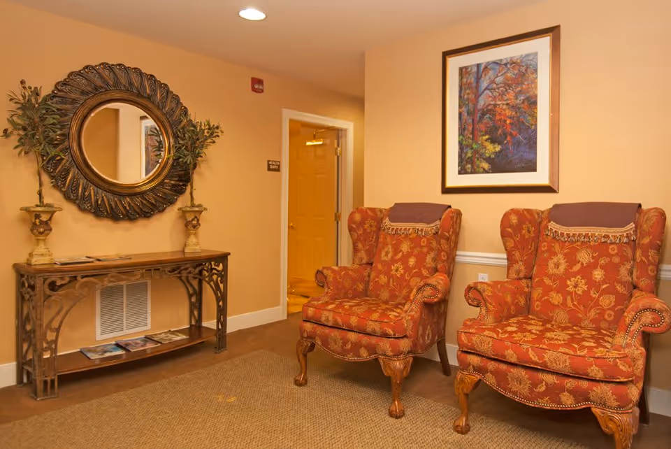 A cozy interior room with two ornate red and gold patterned armchairs placed side by side. On the wall above the chairs is a framed painting of autumn trees. To the left, there is a decorative console table with two small potted plants and a large round mirror with an intricate frame hanging above it. The walls are painted a warm beige color, and there is a door labeled 'Health Suite' in the background.