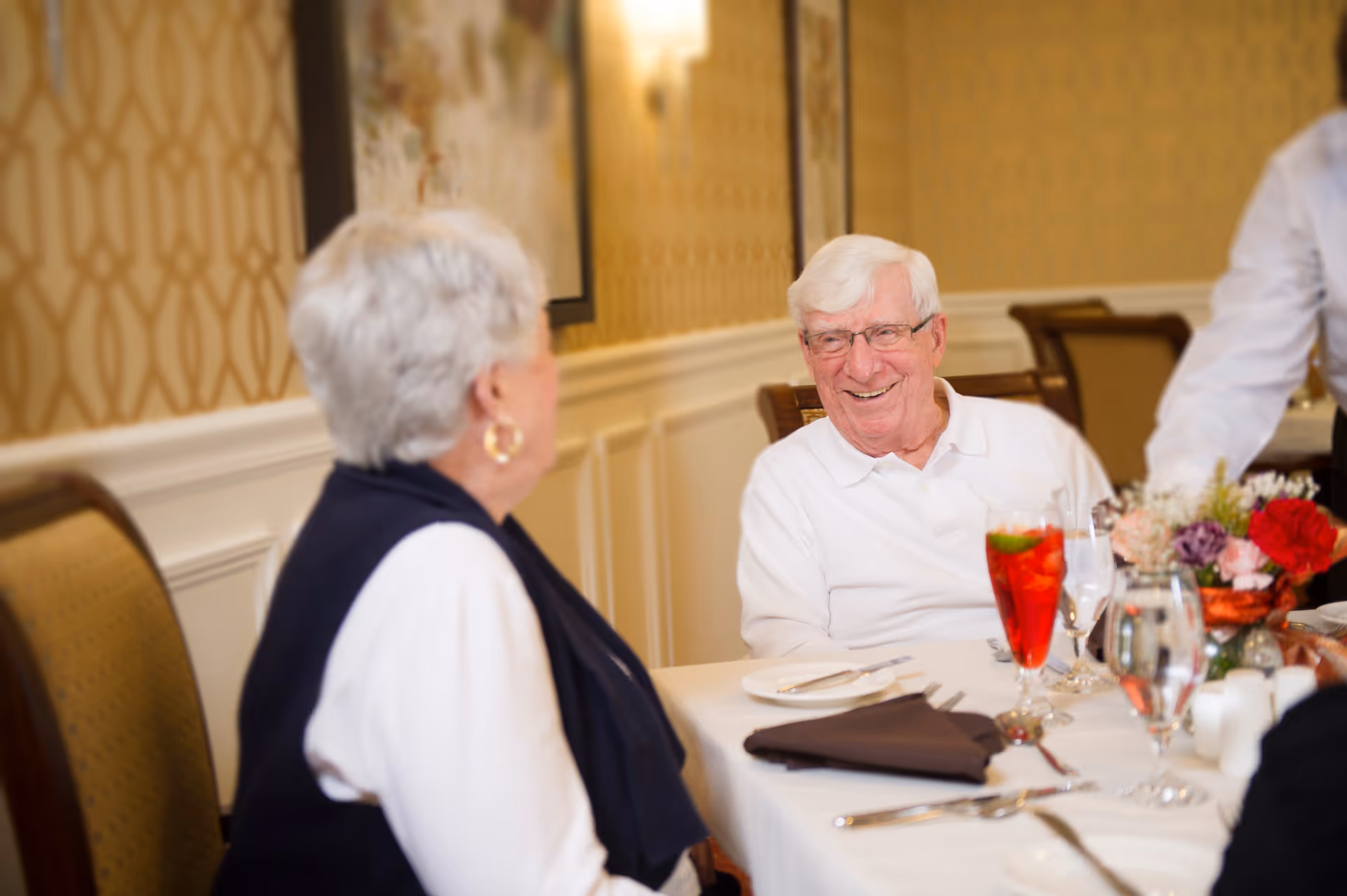 An elderly man and woman sit at a decorated dining table, smiling and conversing in a dining room.