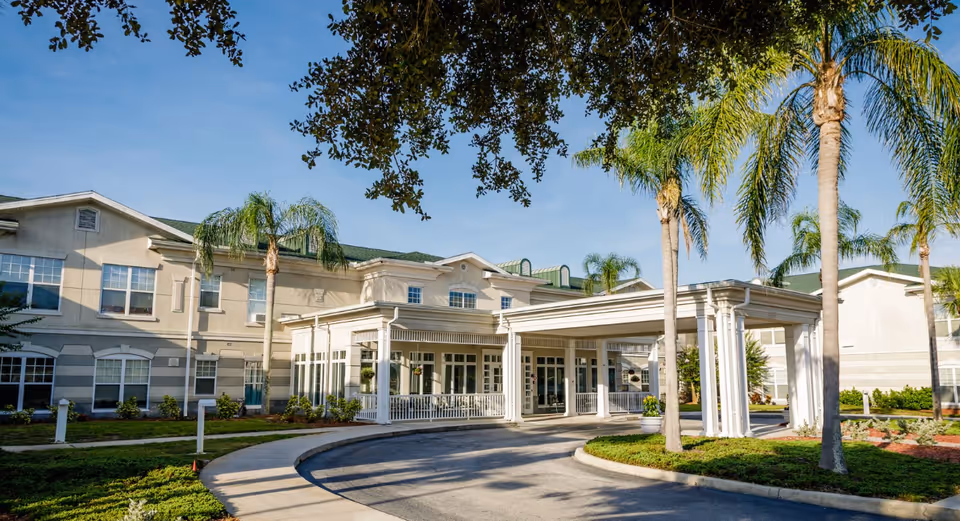 Front entrance of a two-story beige senior living building with a covered porte-cochere, palm trees, and a landscaped circular driveway.