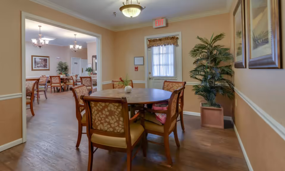 Small dining area with a round wooden table surrounded by upholstered chairs, a potted plant, and a view into a larger dining room.