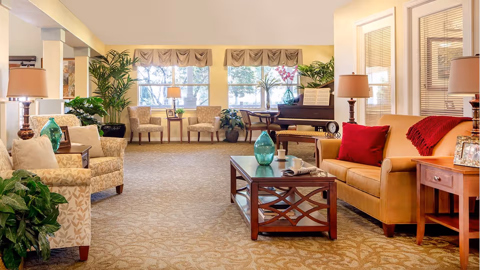 A bright and spacious living room area in a senior living facility with beige patterned armchairs, a tan sofa with red cushions and a red throw, wooden coffee and side tables with lamps, green plants, and a piano near large windows with beige valances letting in natural light.