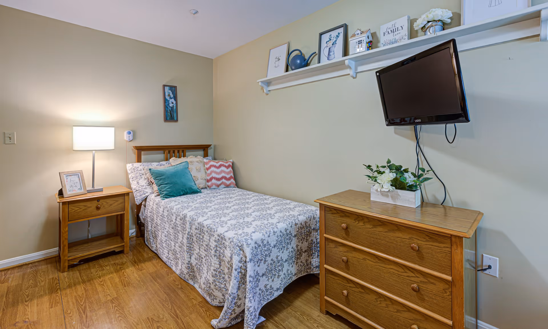 A cozy bedroom with a single bed covered in a patterned bedspread and several decorative pillows. Next to the bed is a wooden nightstand with a lamp and a framed picture. On the opposite wall, there is a wooden dresser with a potted plant on top and a flat-screen TV mounted above it. A shelf above the TV holds framed pictures and decorative items. The room has wooden flooring and light-colored walls.