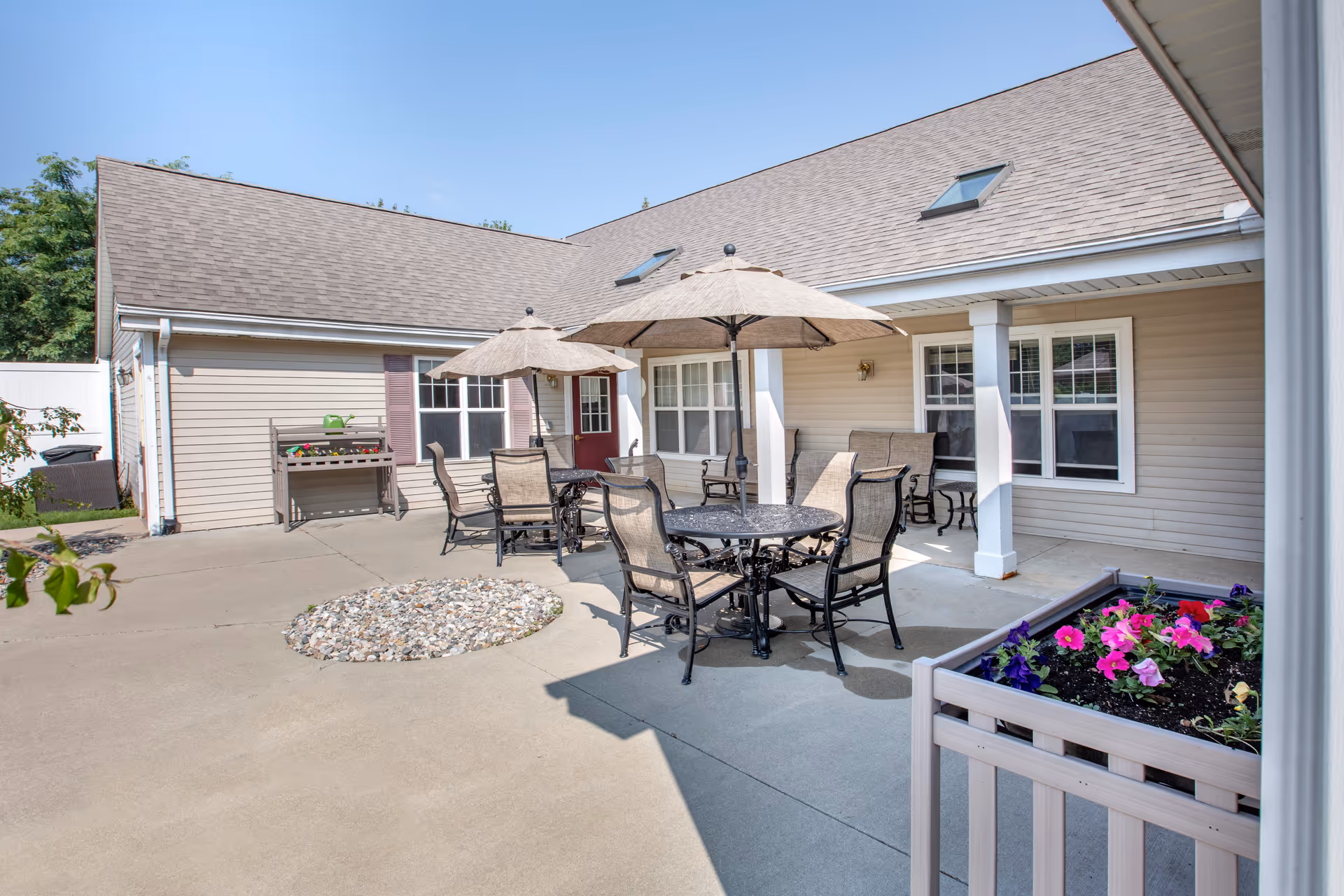 Outdoor patio area at Brookdale Saginaw with round metal tables and chairs under beige umbrellas. The patio is surrounded by beige siding buildings with white trim and windows. There is a flower planter with colorful flowers in the foreground and a small rock garden in the center of the concrete patio.