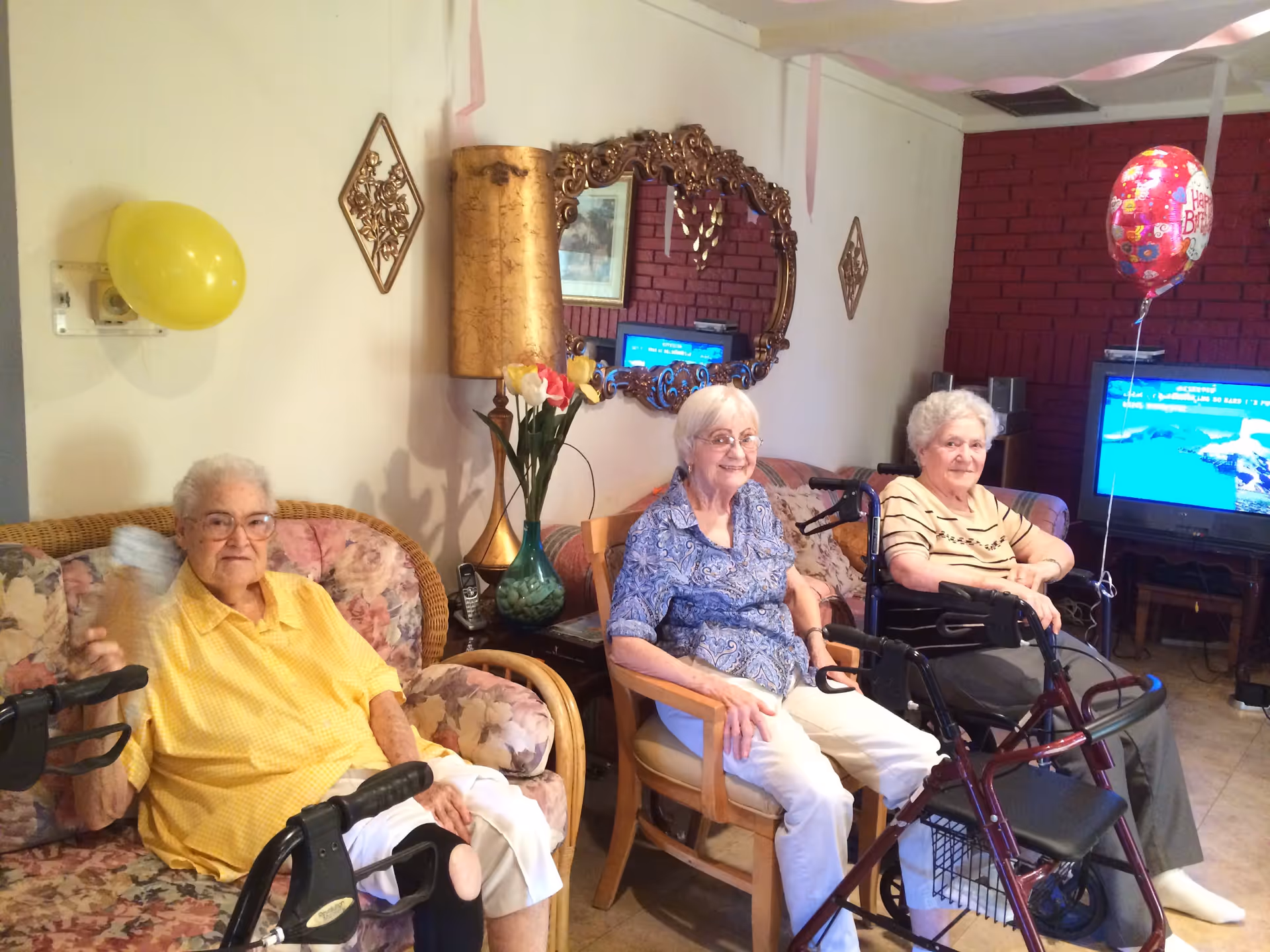 Three elderly women sitting in a living room decorated with balloons and streamers, with a TV and a large ornate mirror on the wall behind them. Two of the women have walkers in front of them.