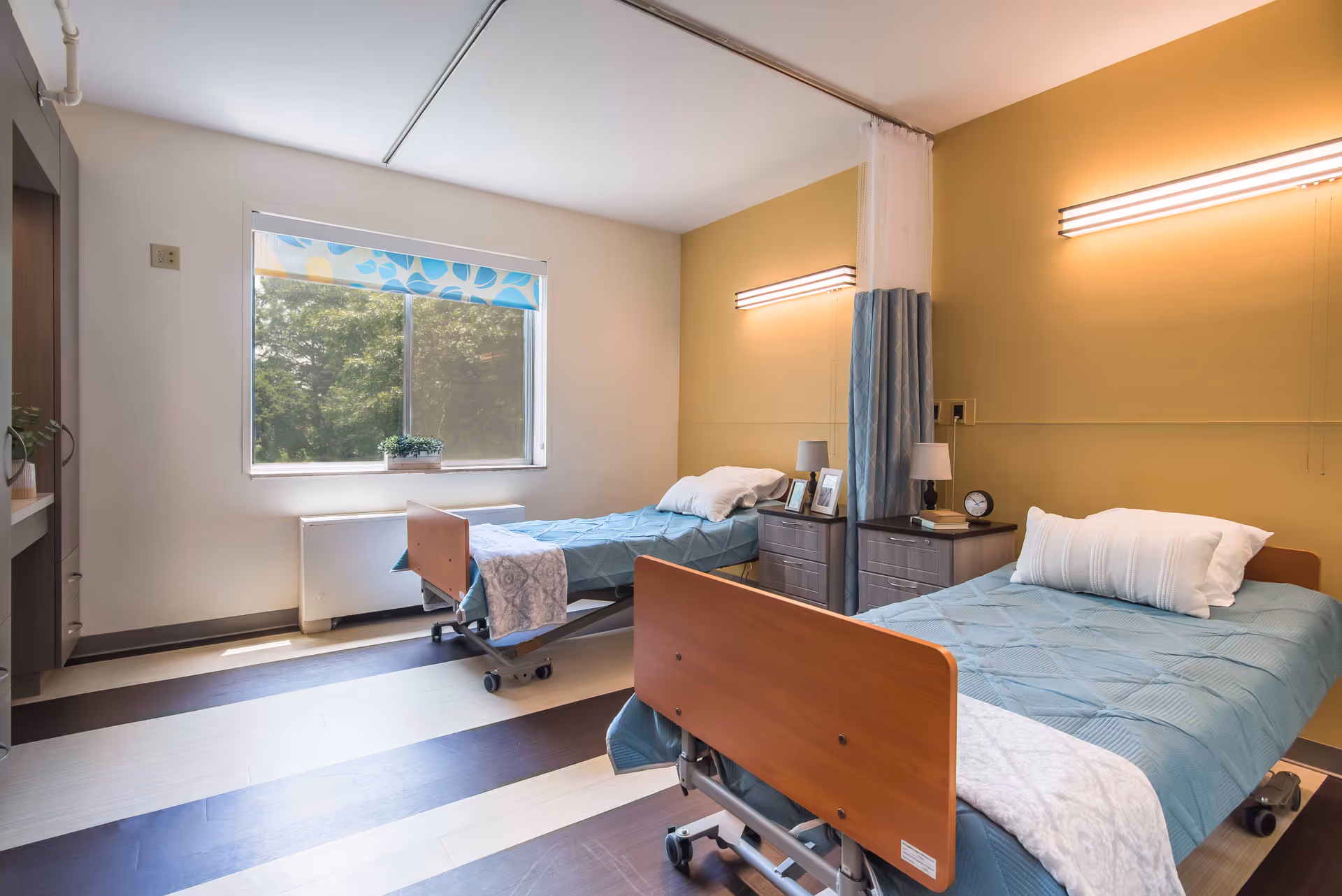 A bright and clean senior living room with two hospital-style beds, each with blue bedding and white pillows. There is a window with a blue patterned shade letting in natural light. Between the beds are two nightstands with lamps, a clock, and framed photos. The walls are painted yellow and white, and there is a privacy curtain between the beds.