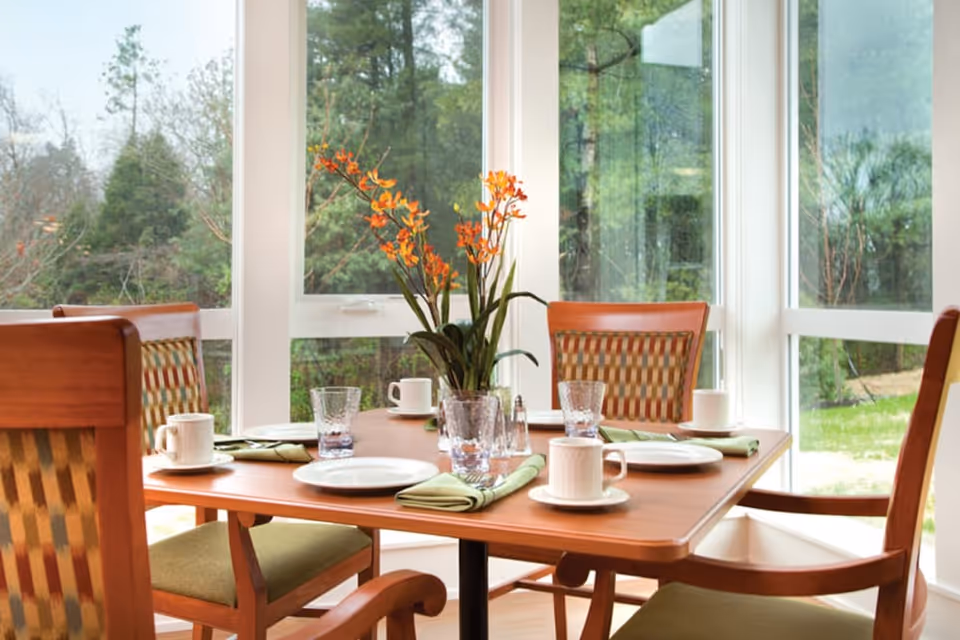 A dining table set for four with white plates, cups, glasses, and green napkins, surrounded by wooden chairs with patterned cushions. A vase with orange flowers is placed in the center of the table. Large windows behind the table show a view of green trees and outdoor greenery.