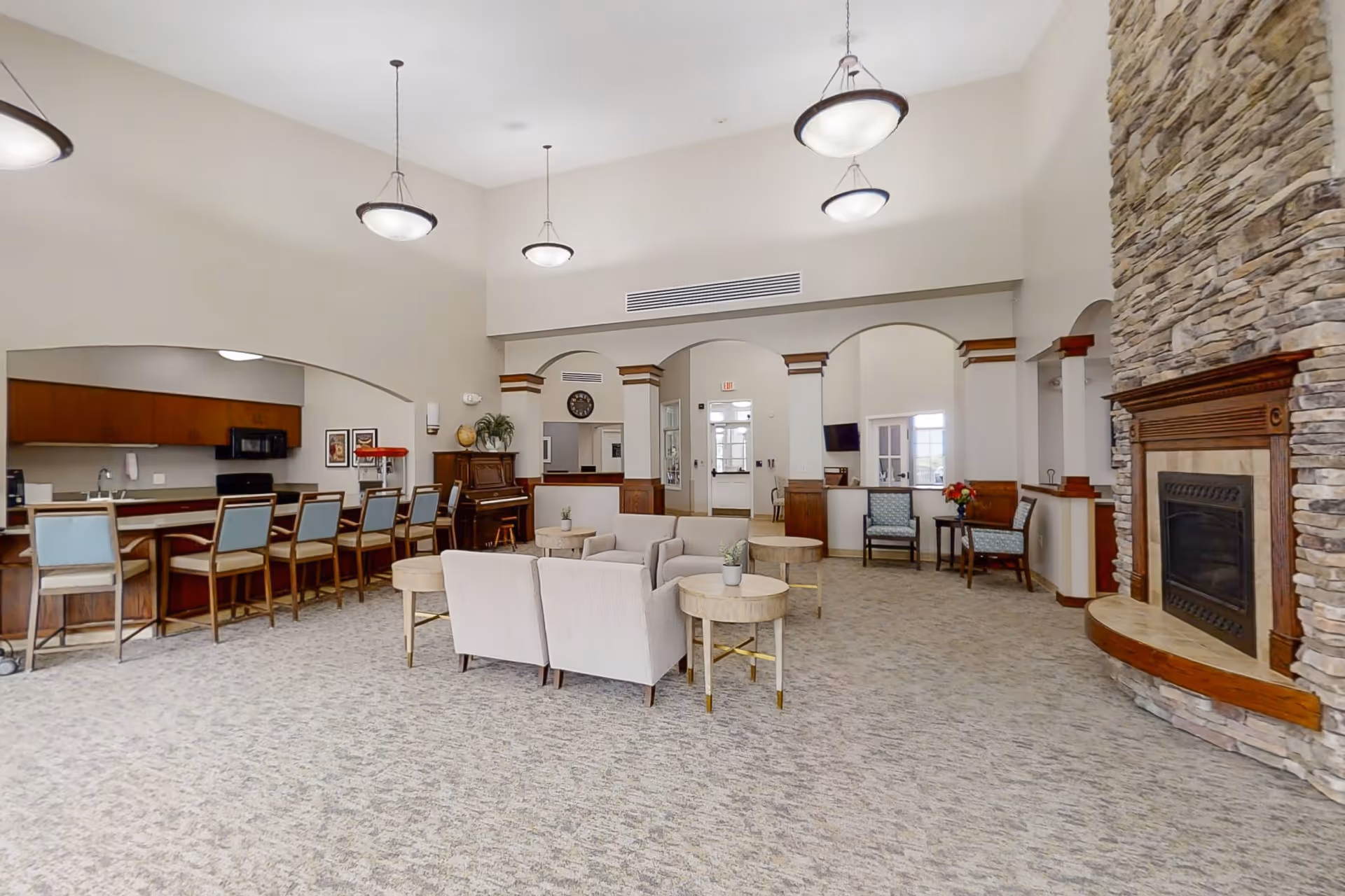 A spacious living room area in CountryHouse Residence featuring a stone fireplace on the right, several beige armchairs arranged around round wooden tables in the center, and a long counter with bar stools on the left. The room has high ceilings with hanging light fixtures and neutral-colored walls and carpet. There is a piano against the back wall and a small seating area with two chairs and a table near the back right corner.