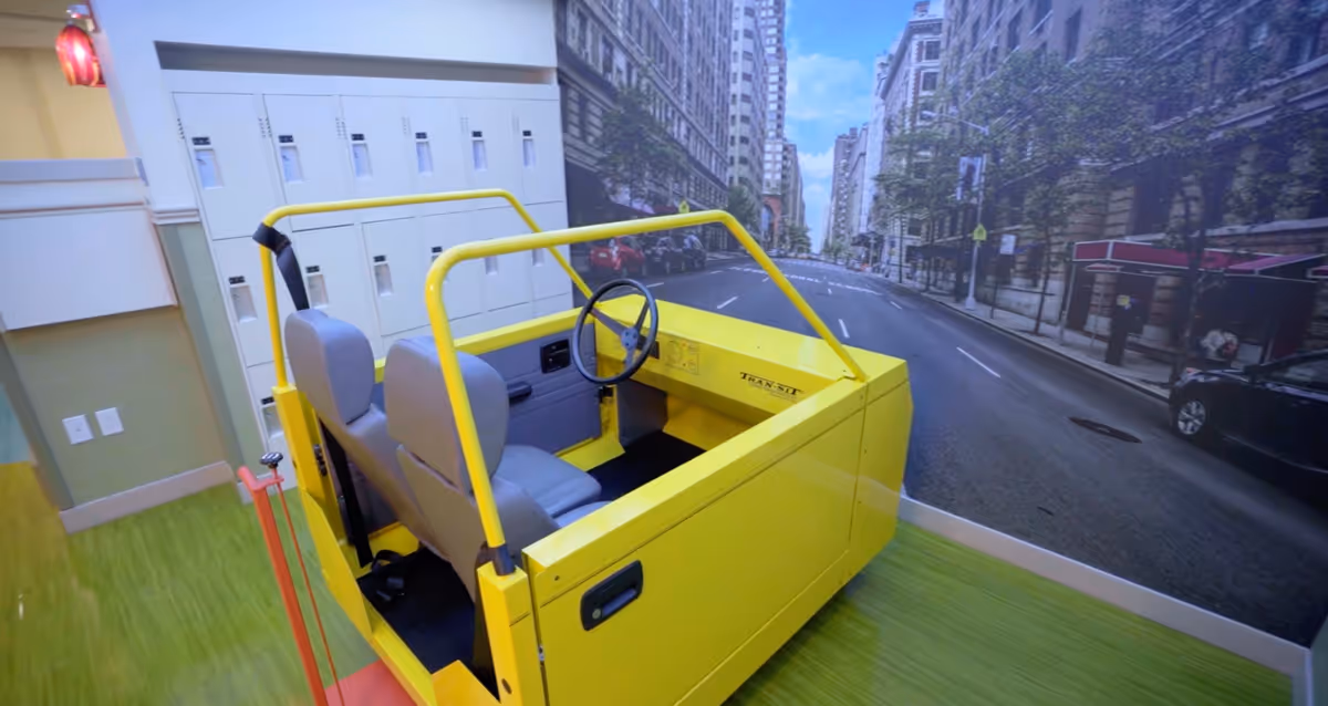 Indoor room with a yellow vehicle frame resembling a small car or driving simulator with two gray seats and a steering wheel, placed on a green floor. The background features a large wall mural depicting a city street with tall buildings and trees.