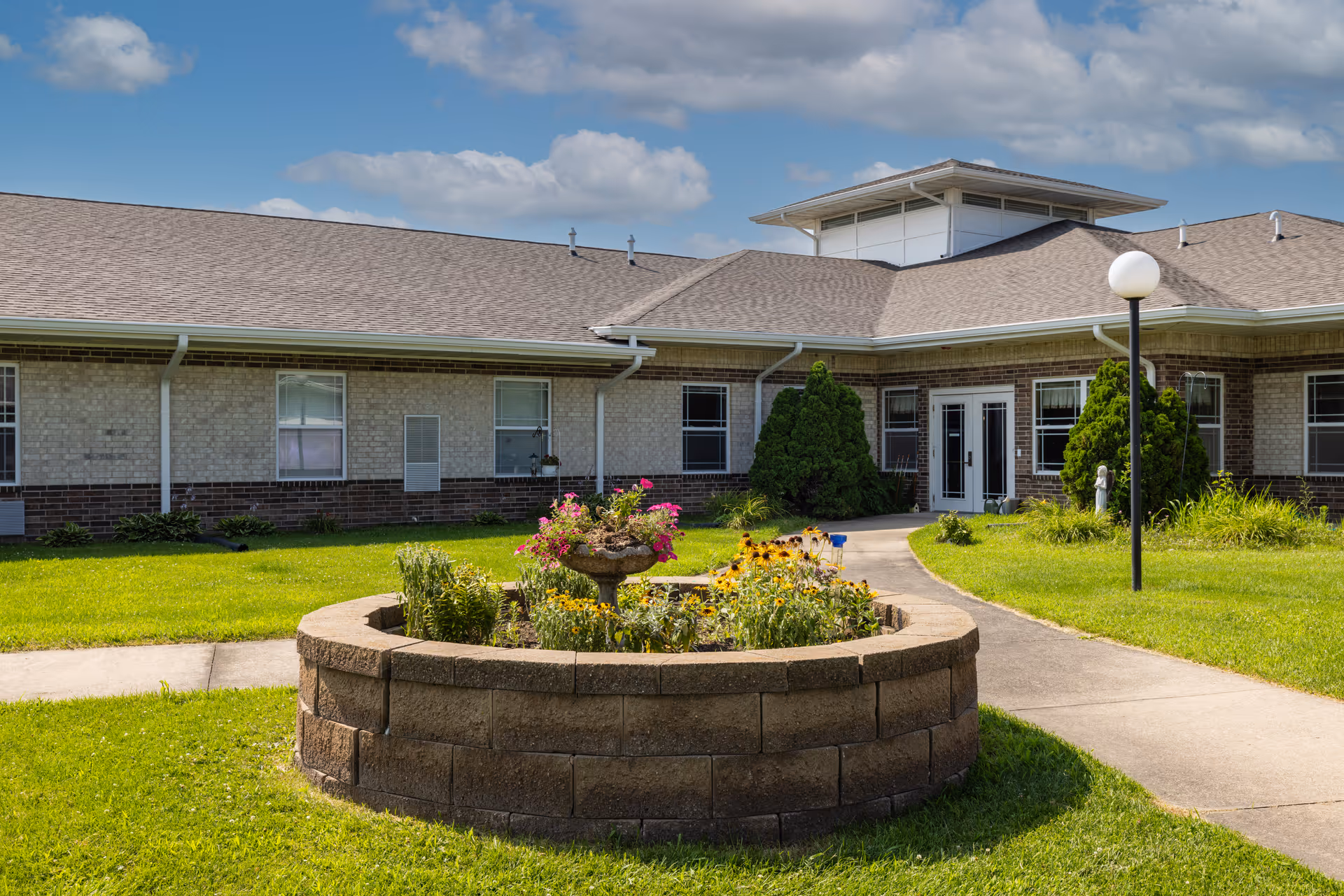 Exterior front of a single-story senior living facility with a circular raised flower bed and walkway to the entrance.
