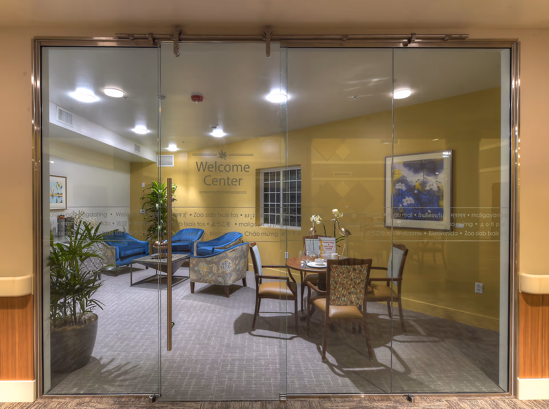 View through glass doors into a welcome center with a seating area featuring blue and patterned chairs around a coffee table, a round table with four chairs, potted plants, and a framed artwork on a yellow wall.