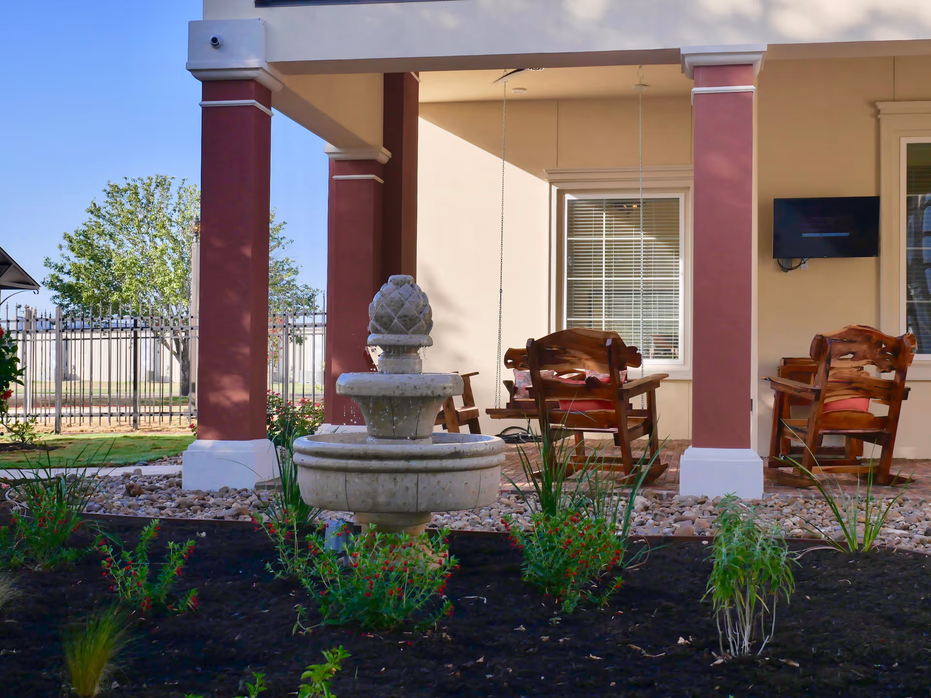 Outdoor patio area with a stone water fountain in the foreground, surrounded by small plants and flowers. The patio has two large columns painted in beige and maroon, wooden chairs with red cushions, a hanging wooden swing, and a wall-mounted TV. A tree and a metal fence are visible in the background under a clear blue sky.