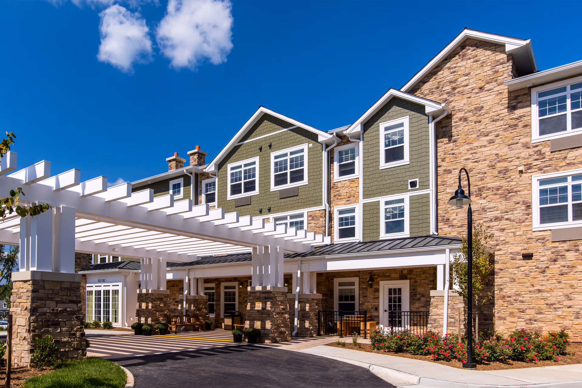 Exterior view of a multi-story assisted living facility with stone and green siding, white trim, and a covered entrance with white pergola beams. There are flower beds and a black street lamp near the entrance under a bright blue sky with a few clouds.