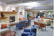 A spacious and well-lit living room area with multiple seating options including sofas, armchairs, and small tables. There is a fireplace on the left wall with decorative shelves above it, and a chandelier hanging from the ceiling. The room has neutral-colored walls and carpeted floors, creating a comfortable and inviting atmosphere.