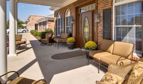 A covered outdoor patio area with cushioned chairs and small tables, potted yellow flowers, and a brick exterior wall with windows and a door featuring an oval glass panel.