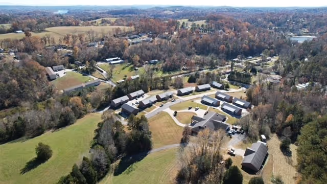 Aerial view of a senior living facility named Small Town Senior Living at Deer Ridge, showing multiple single-story buildings arranged in a curved layout surrounded by trees and open grassy areas, with a rural landscape in the background.