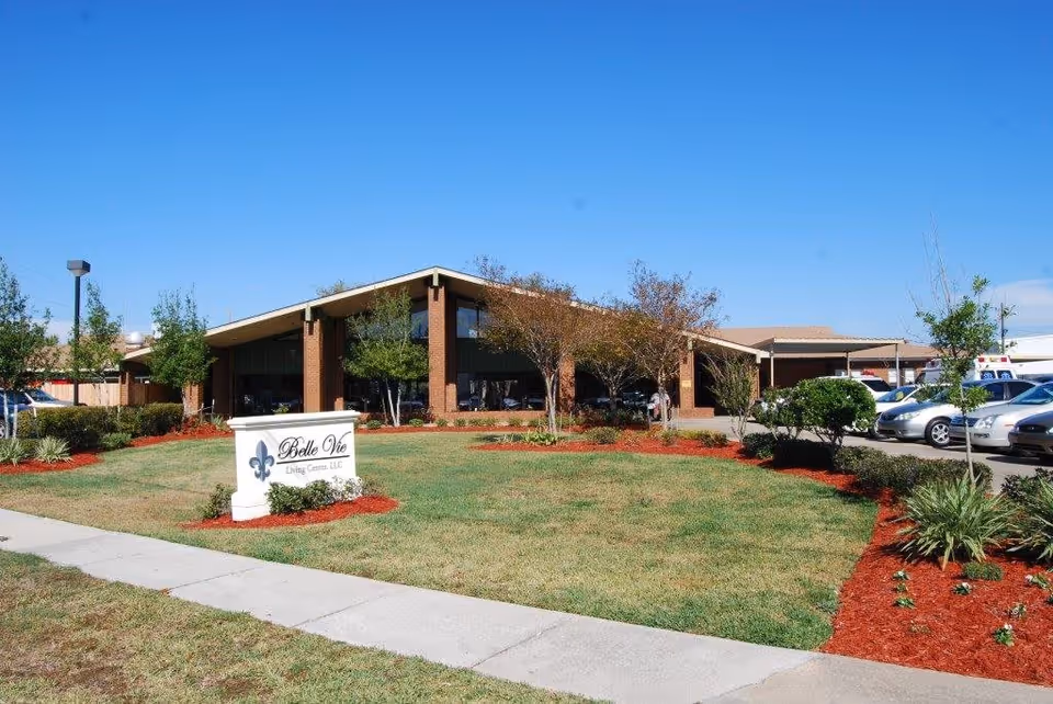 Exterior view of Belle Vie Living Center, LLC, a single-story brick building with a slanted roof, surrounded by a well-maintained lawn, small trees, and landscaped flower beds with red mulch. Several cars are parked in the parking lot to the right of the building under a clear blue sky.