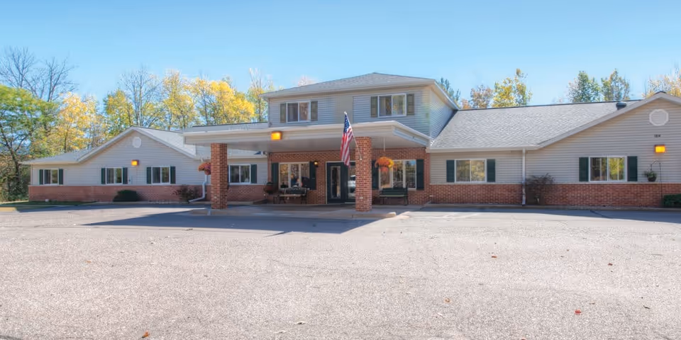 Single-story assisted living building with a covered entrance, American flag, and a large parking area in front.