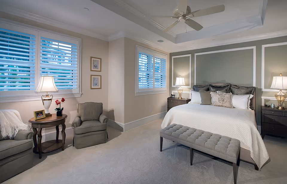 Spacious, elegantly decorated bedroom with a large bed, tufted bench, nightstands with lamps, and seating by shuttered windows.
