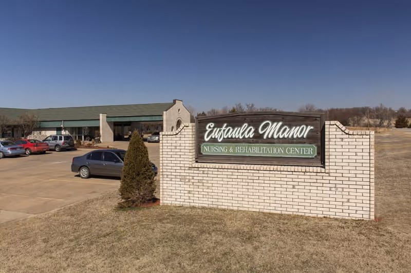Brick sign reading "Eufaula Manor Nursing & Rehabilitation Center" in front of a single-story nursing facility with parked cars.