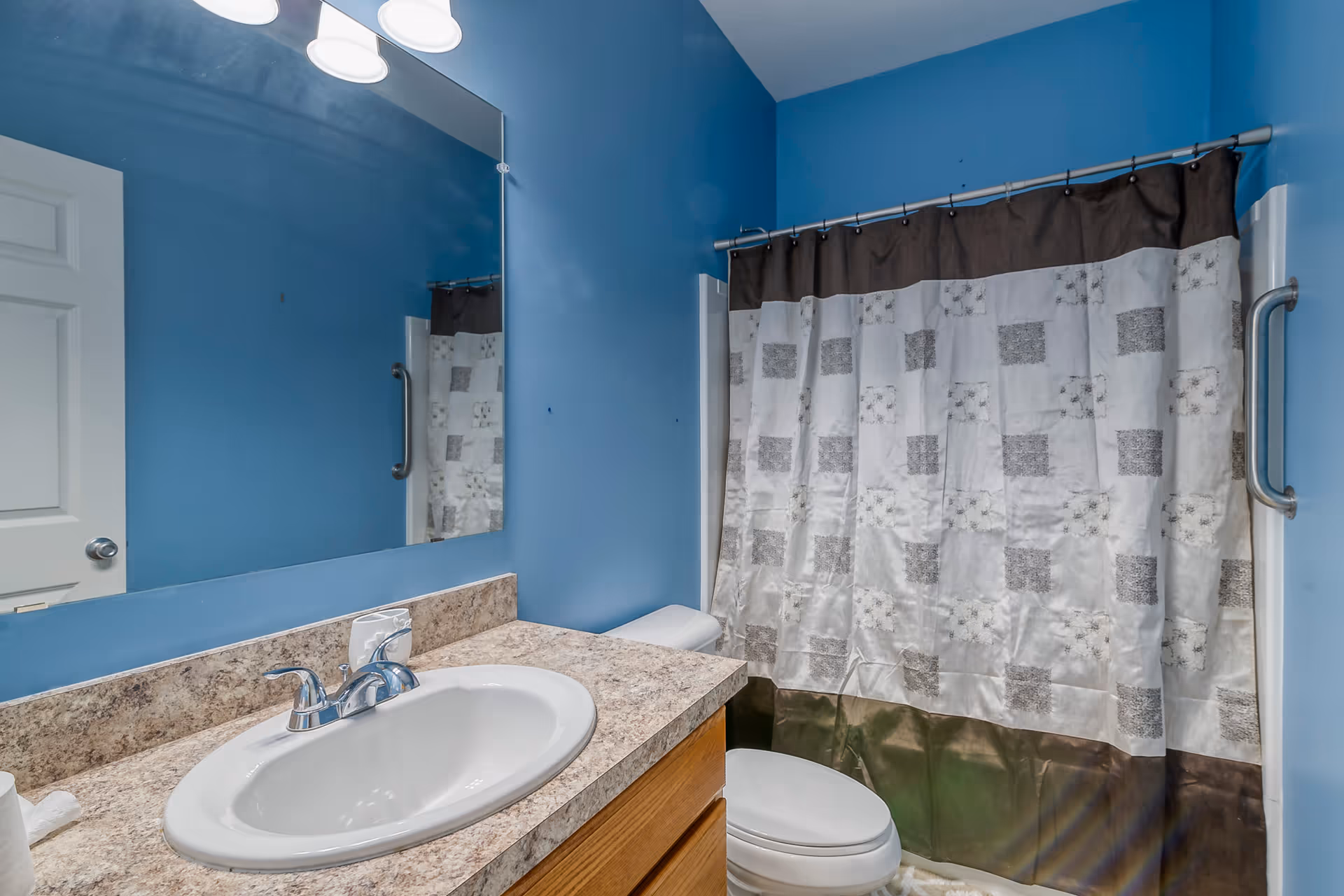 Small bathroom with blue walls, a sink and countertop, mirror, toilet, and a shower with a patterned curtain.