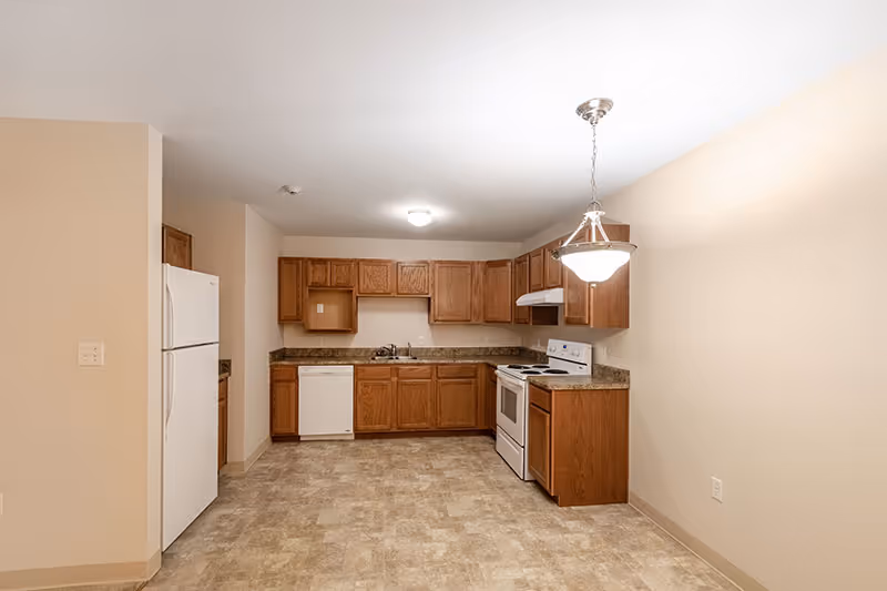 A clean and empty kitchen with wooden cabinets, a white refrigerator, a dishwasher, an electric stove with an oven, and a hanging light fixture. The floor is covered with beige tiles and the walls are painted a light cream color.