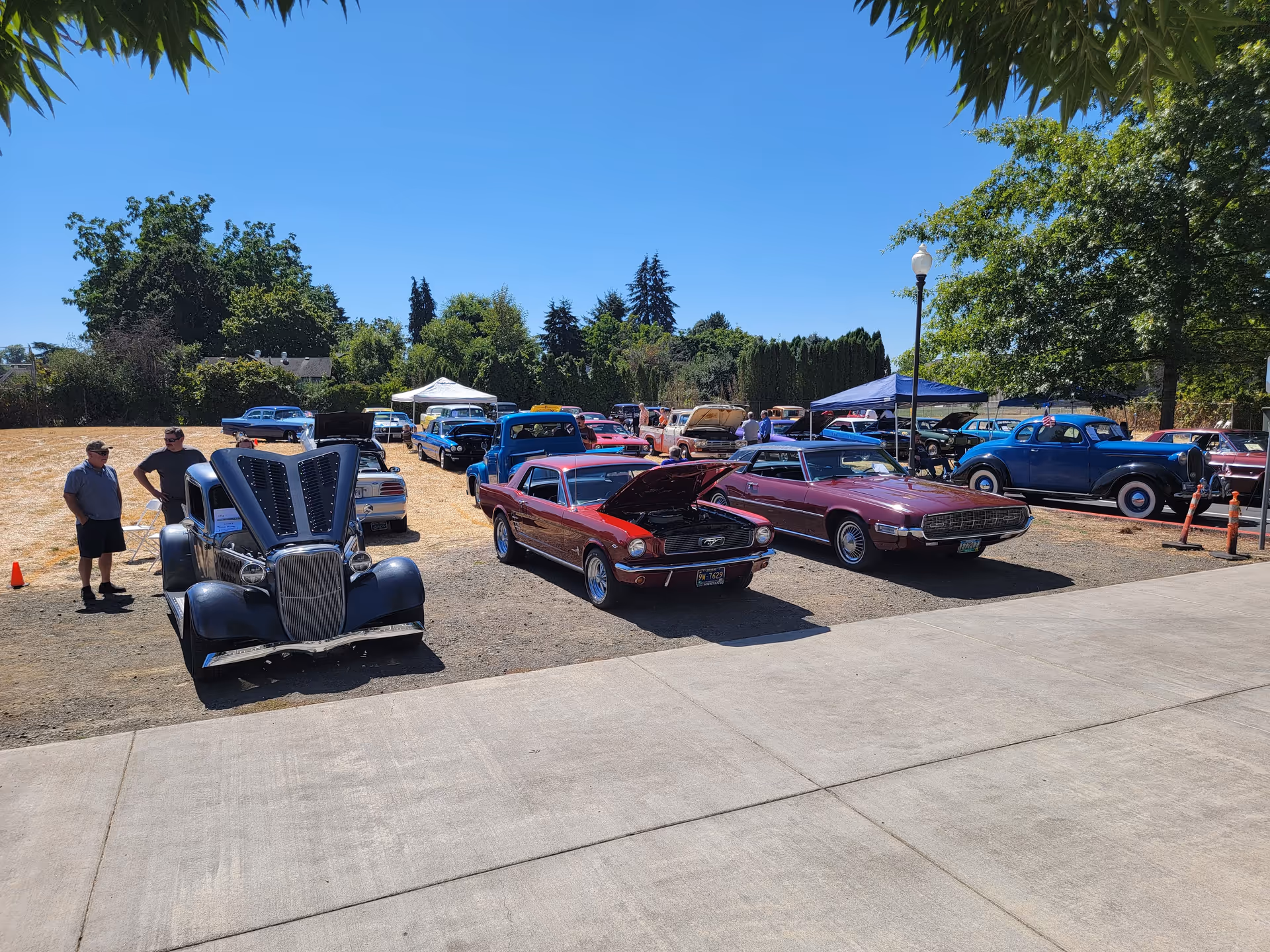A sunny outdoor car show featuring several classic and vintage cars parked on a dirt lot with some people standing nearby. Trees and a clear blue sky are visible in the background.