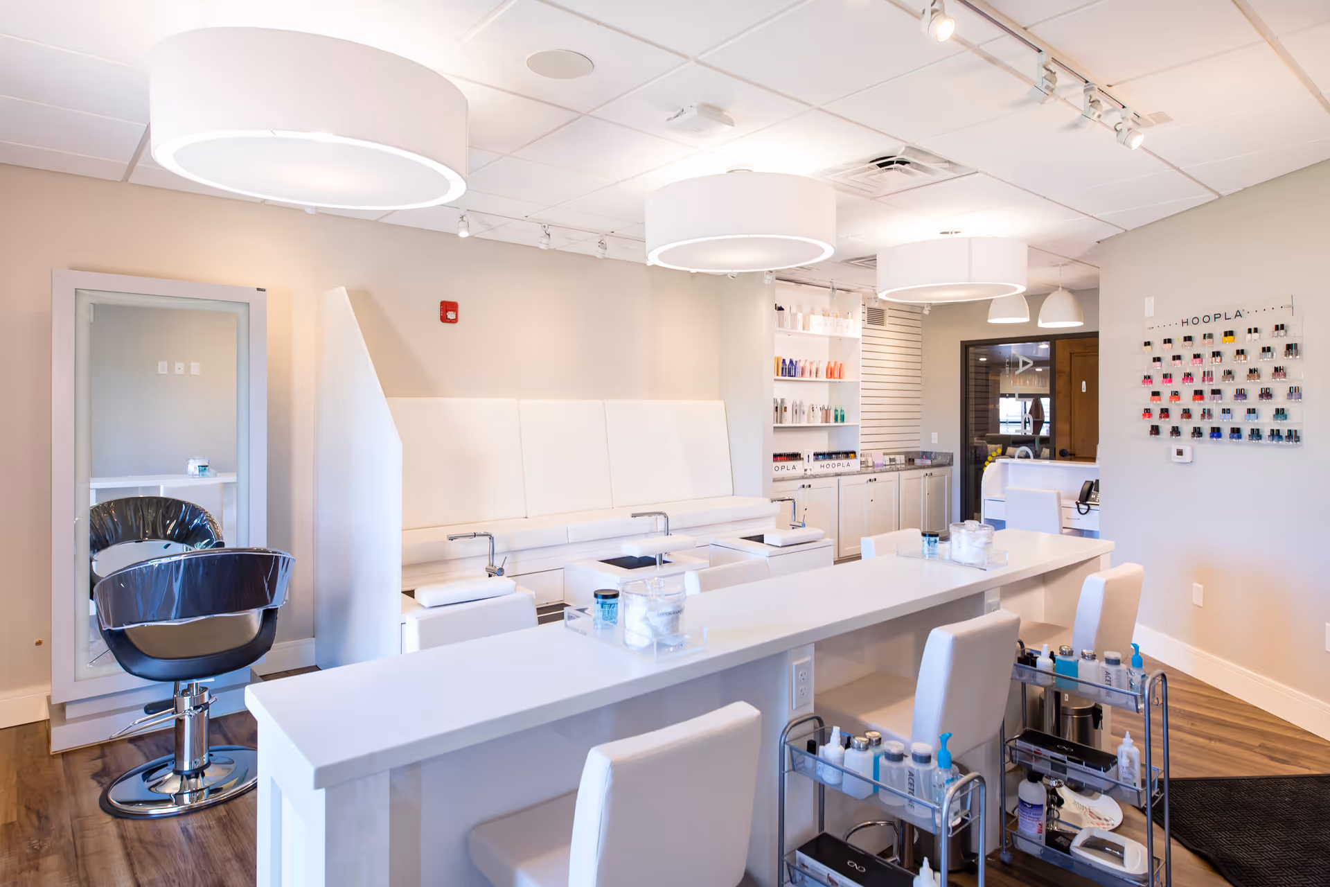 A bright and modern salon area with white furniture including a long table with chairs, manicure stations, and a black salon chair in front of a large mirror. Shelves with beauty products and a nail polish display are visible on the walls. The room has wooden flooring and large circular ceiling lights.