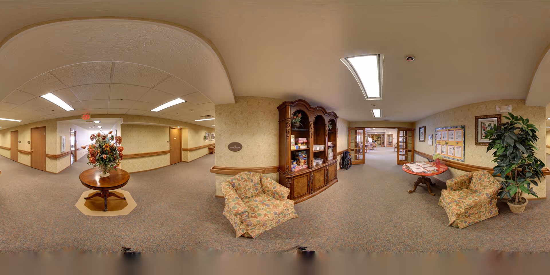 A spacious and well-lit hallway in a senior living facility with beige walls and carpeted floors. There is a round wooden table with a large floral arrangement in the center. Along the walls are comfortable floral-patterned armchairs, a wooden cabinet with shelves holding board games and other items, a bulletin board with notices, and a potted plant. The hallway leads to other rooms and areas, with wooden doors and framed artwork on the walls.