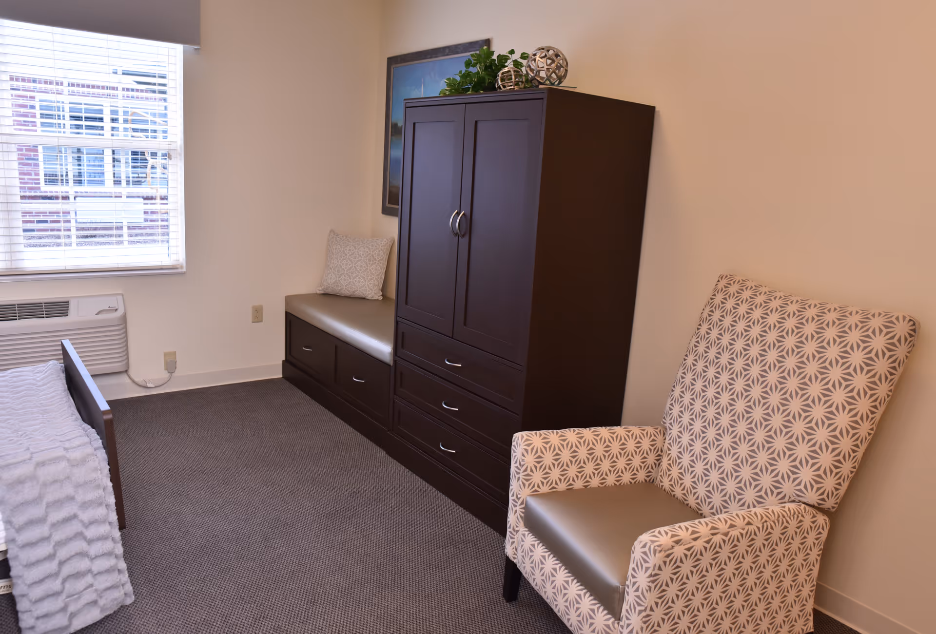 A small, neatly arranged bedroom with a single bed partially visible on the left, a window with blinds, a cushioned bench with a decorative pillow, a dark wooden wardrobe with drawers, and a patterned armchair on the right.