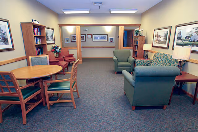A cozy living room area with a round wooden table and four chairs on the left, two red armchairs near a bookshelf with books and a flower arrangement, and a green armchair and a green patterned sofa on the right. The walls are decorated with framed pictures and there is a lamp on a side table next to the sofa. The room has carpeted flooring and soft overhead lighting.