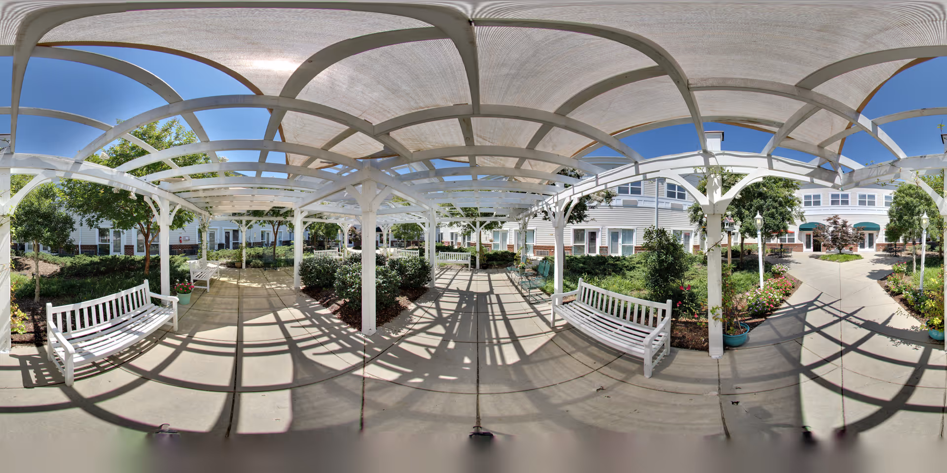 Outdoor garden area at Morningview at Irving Park featuring a white pergola with a translucent roof casting shadows on the concrete walkway. White benches are placed along the path, surrounded by green shrubs, potted plants, and trees. The background shows a multi-story building with windows and green awnings under a clear blue sky.