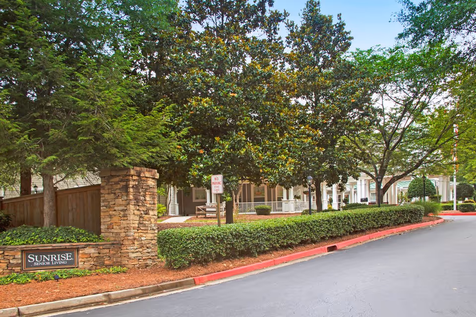 Landscaped driveway and entrance to a senior living facility with a stone sign reading 'SUNRISE SENIOR LIVING' and a porch-front building partially hidden by trees.