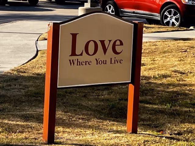 A beige sign with red wooden posts on a grassy area near a sidewalk and parked cars. The sign reads 'Love Where You Live'.