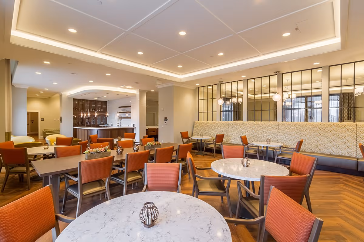 A spacious and well-lit dining room with multiple round and rectangular tables surrounded by orange cushioned chairs. The room features a modern ceiling with recessed lighting, a wooden floor, and a long cushioned bench along one wall with decorative mirrors above it. In the background, there is a bar area with pendant lights and shelves.