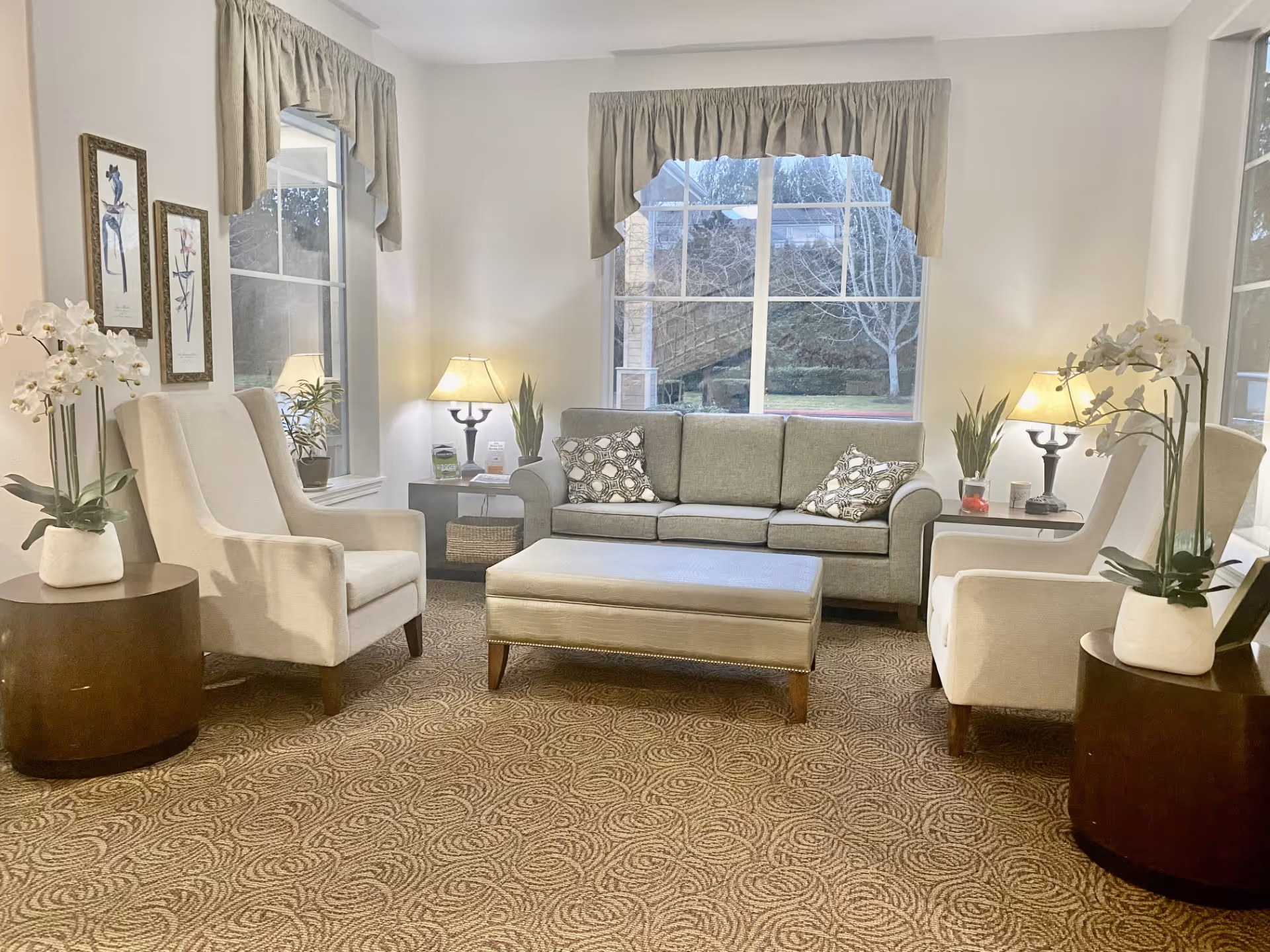 A cozy seating area with a sofa, two armchairs, an ottoman and table lamps in a bright living room with large windows.