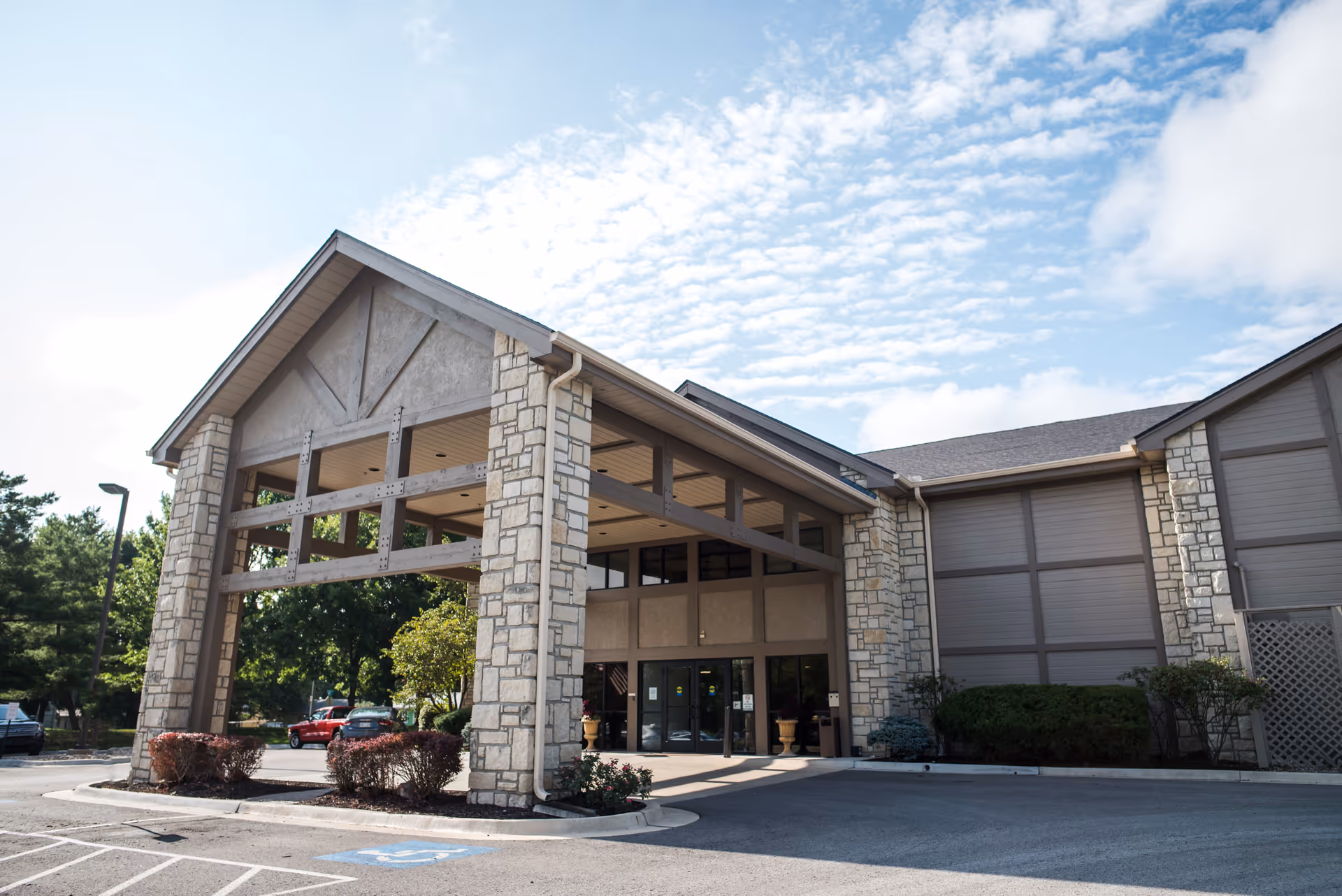 Front entrance of a healthcare building with a large covered portico supported by stone pillars and adjacent parking.