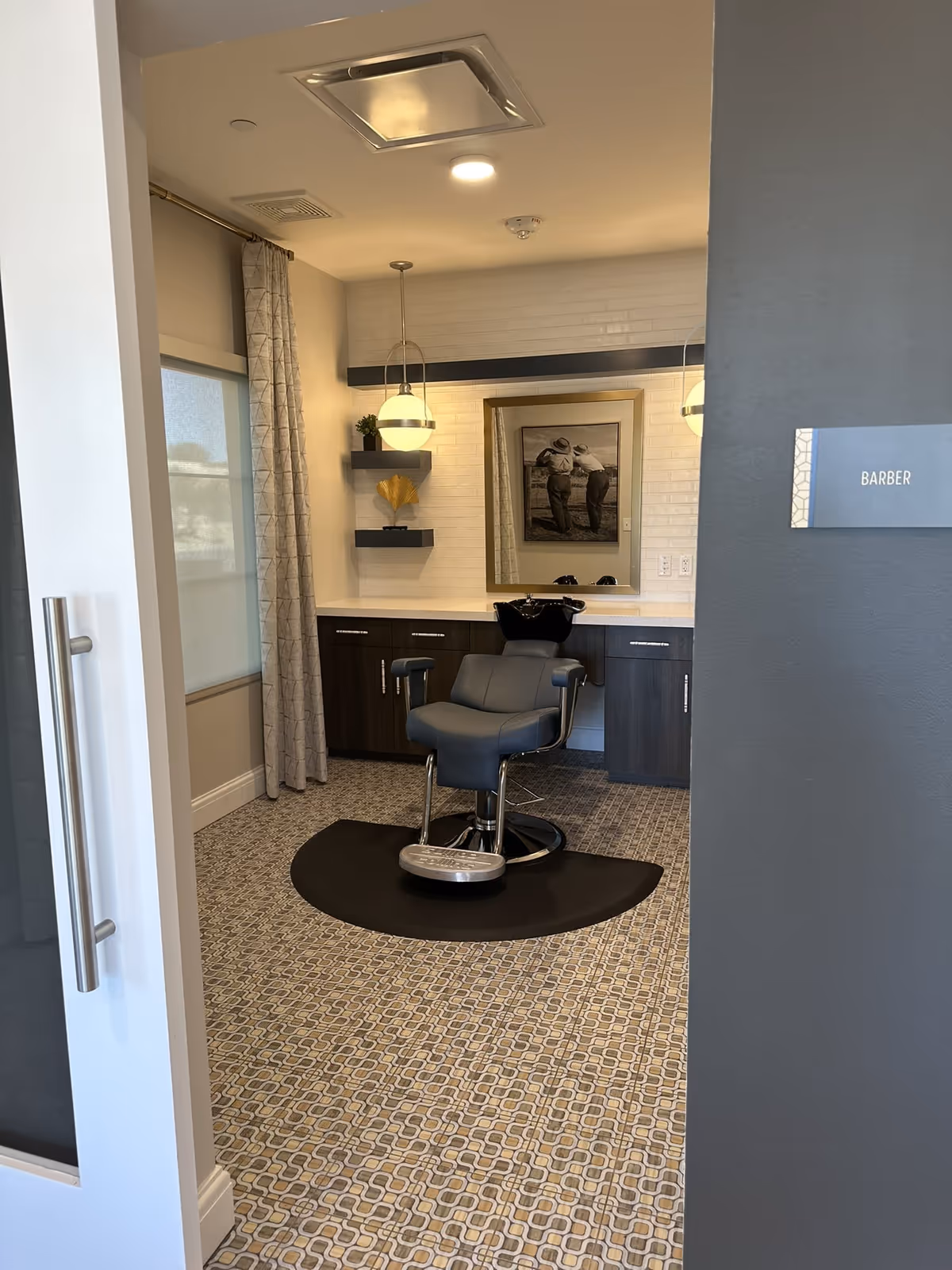 Interior barber room with a salon chair on a mat facing a sink and mirror, framed photo on the wall and a 'BARBER' sign by the doorway.