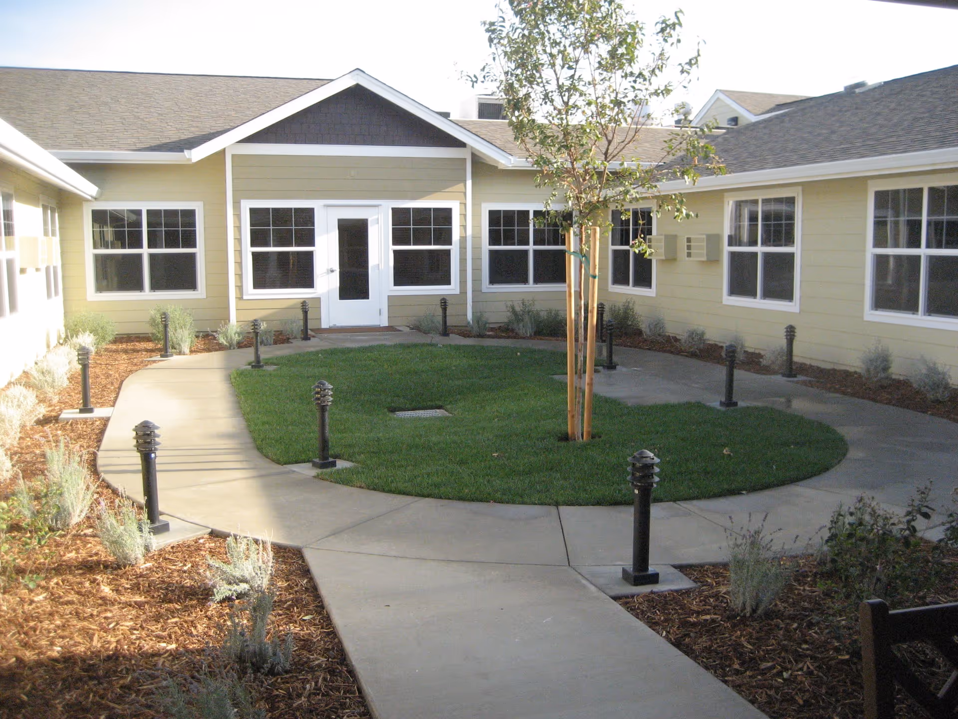 A courtyard area with a circular concrete walkway surrounding a small grassy patch with a young tree in the center. The courtyard is enclosed by a single-story building with beige siding, white trim, and multiple windows. There are small black outdoor lights lining the walkway and some landscaping with mulch and small plants around the edges.