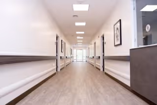 A long, well-lit hallway in a senior living facility with light-colored walls, wooden handrails on both sides, framed artwork on the walls, and a reception desk visible on the right side near the foreground. The hallway has a light wood floor and ceiling lights, leading to a glass door at the far end.