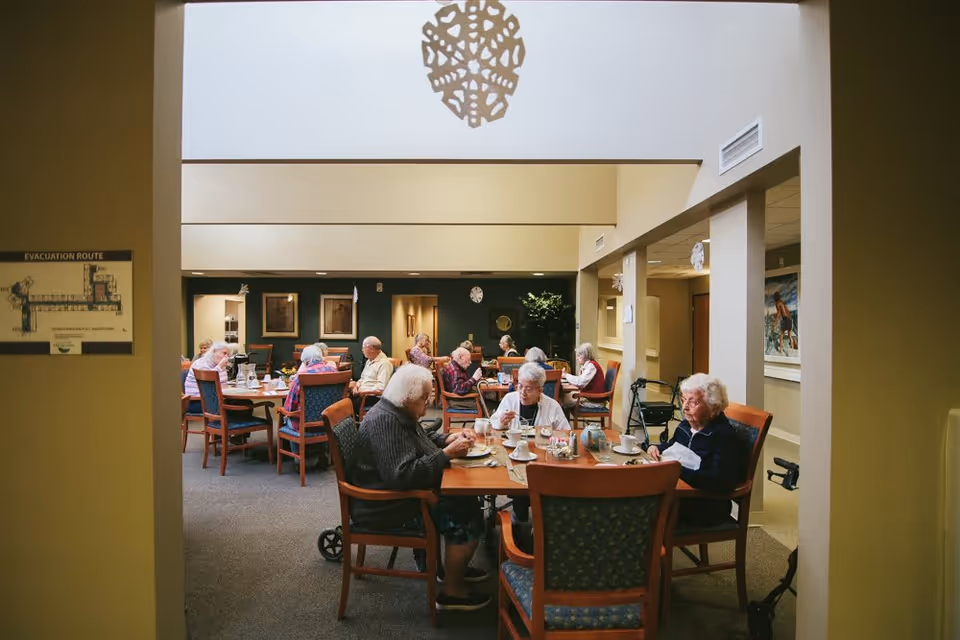 A group of elderly people sitting at tables in a well-lit dining area, eating and socializing. The room has high ceilings, beige walls, and framed artwork on the walls. Some walkers are visible near the tables.