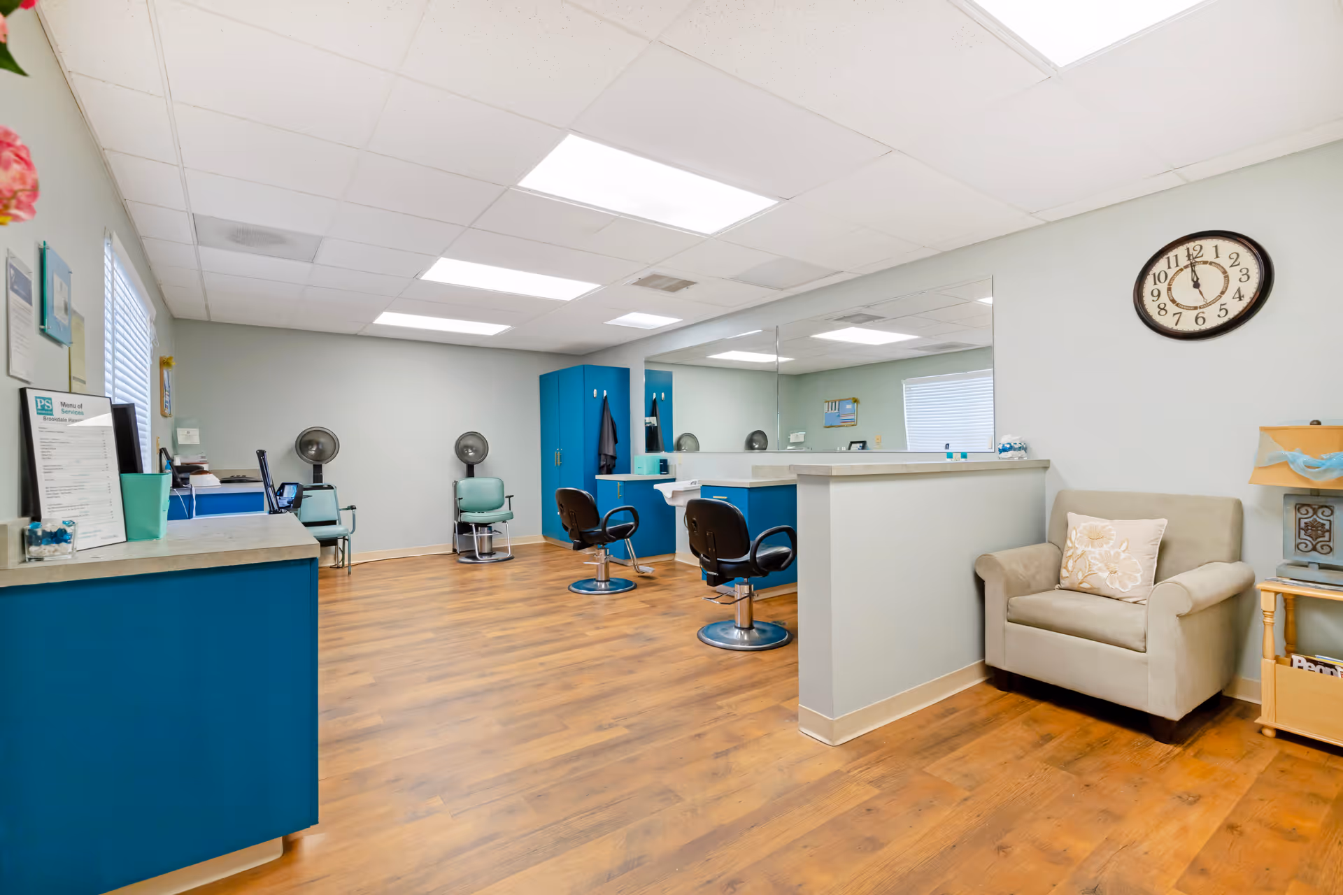 Bright salon-style room with styling chairs, hooded hair dryers, a reception counter, an armchair and a wall clock.