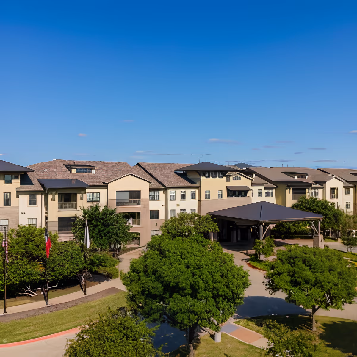 Front exterior of a multi-story senior living building with a covered porte-cochère, trees, and a clear blue sky.