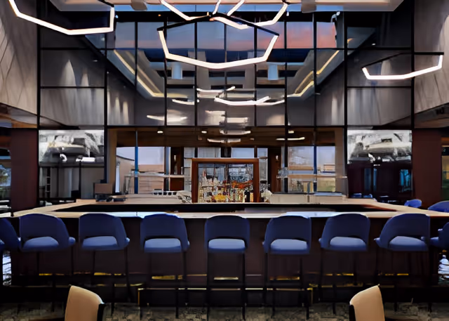 Modern indoor bar area with a long counter and eight blue bar stools. The back wall features large reflective glass panels and shelves with bottles and glassware. Contemporary ceiling lights with geometric designs hang above the bar.