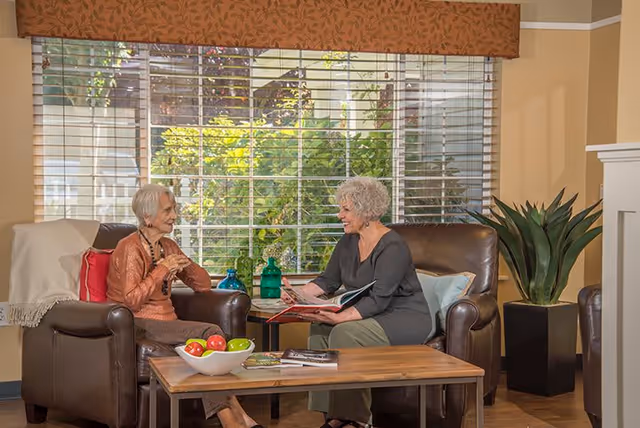 Two women seated in armchairs in a cozy living room, talking and looking at a book across a coffee table with decorative items.