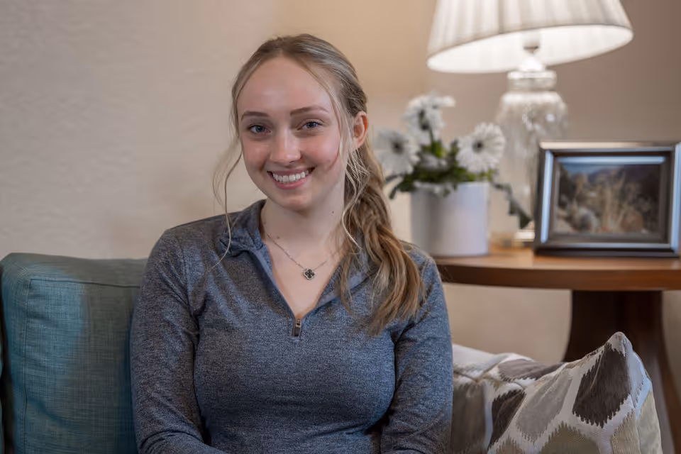 A young woman with blonde hair tied back is sitting on a teal couch in a cozy living room. She is smiling and wearing a gray zip-up top. Behind her is a wooden side table with a white vase of white flowers, a framed picture, and a glass lamp with a white lampshade. There is also a patterned cushion on the couch.
