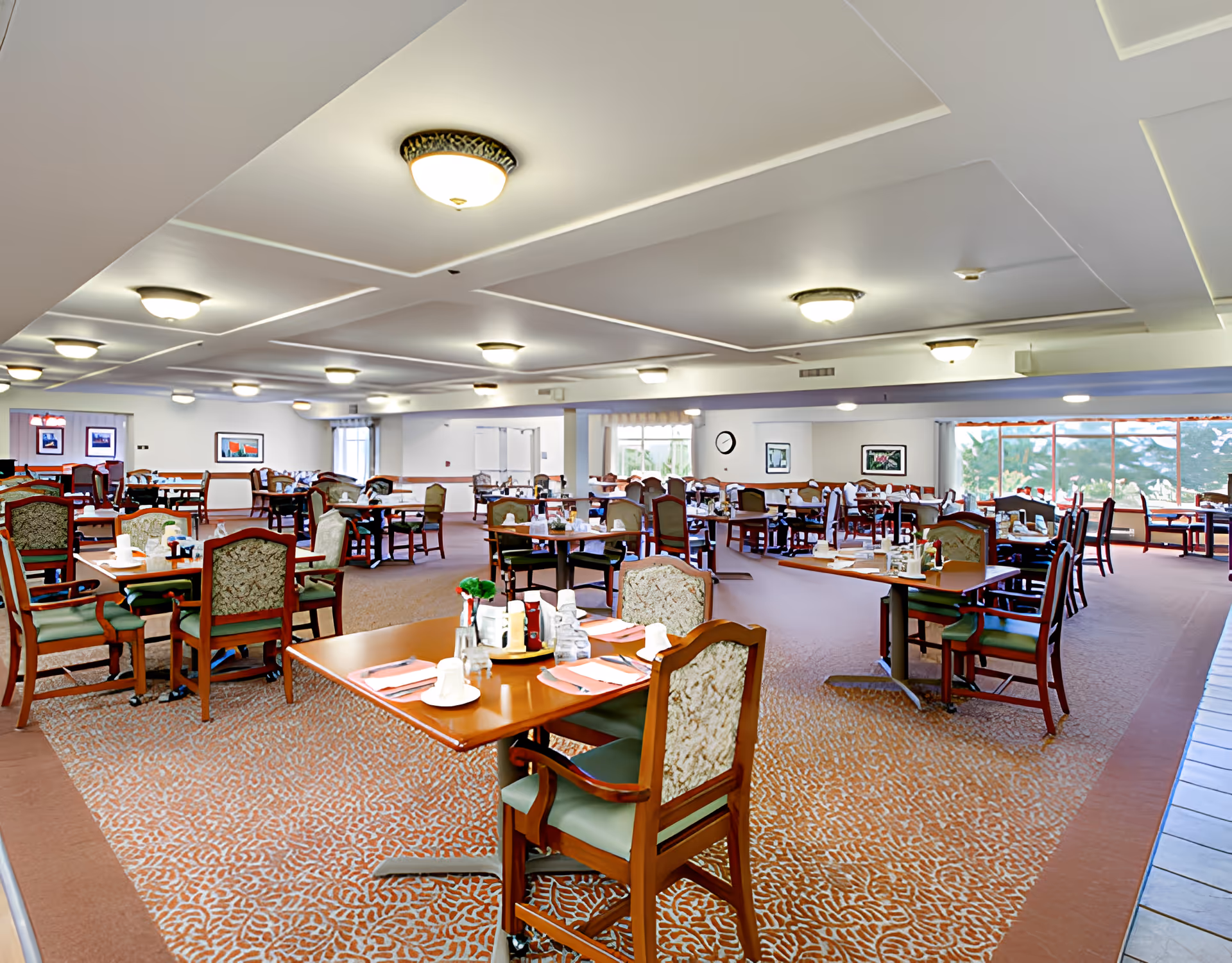 A spacious dining room in a senior living facility with multiple wooden tables and chairs arranged neatly. Each table is set with placemats, napkins, cups, and condiments. The room has patterned carpet flooring, large windows allowing natural light, and ceiling lights providing illumination. Artwork is displayed on the walls.