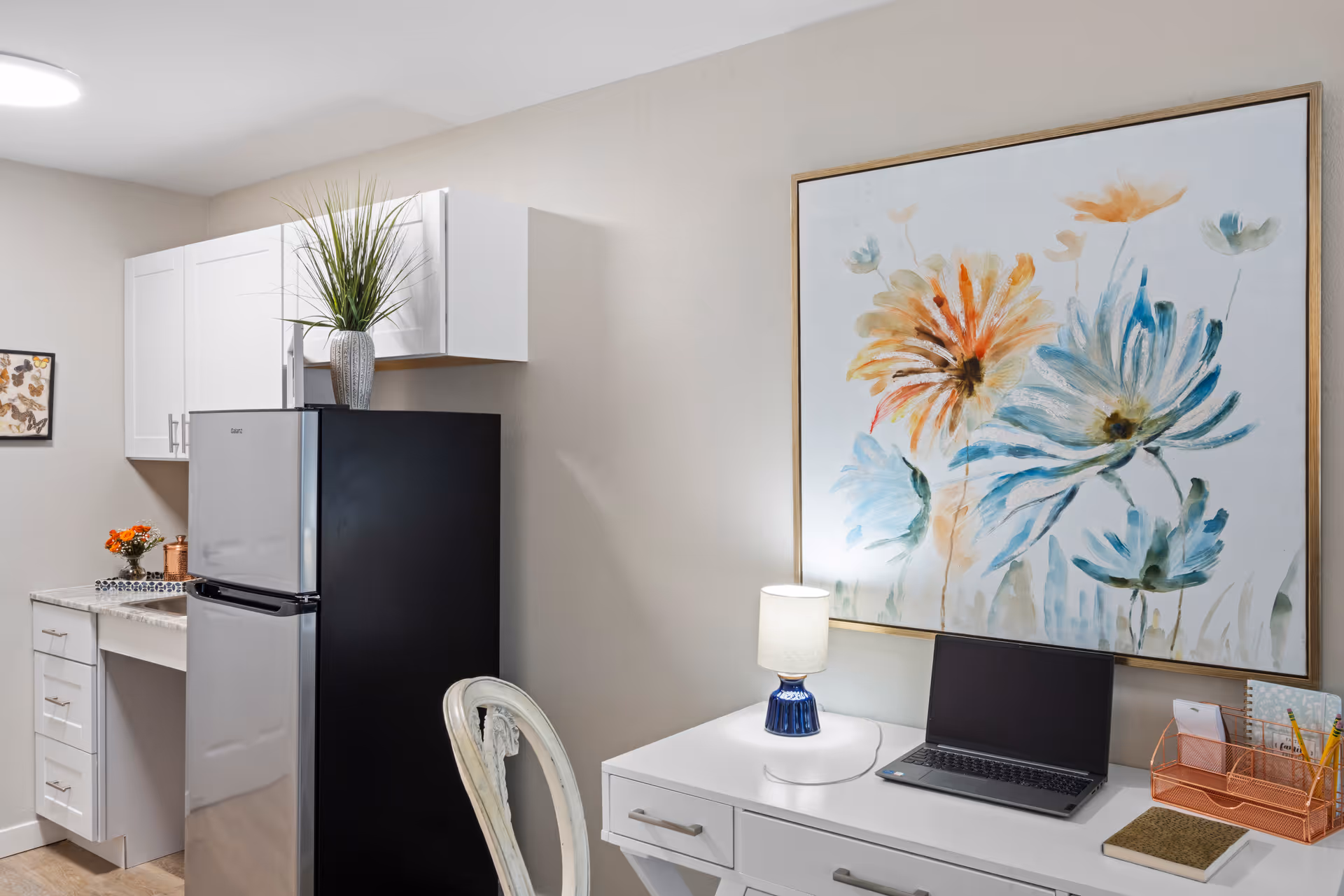 Small kitchenette and workspace featuring a refrigerator and white cabinets next to a desk with a laptop, lamp, and large floral painting.