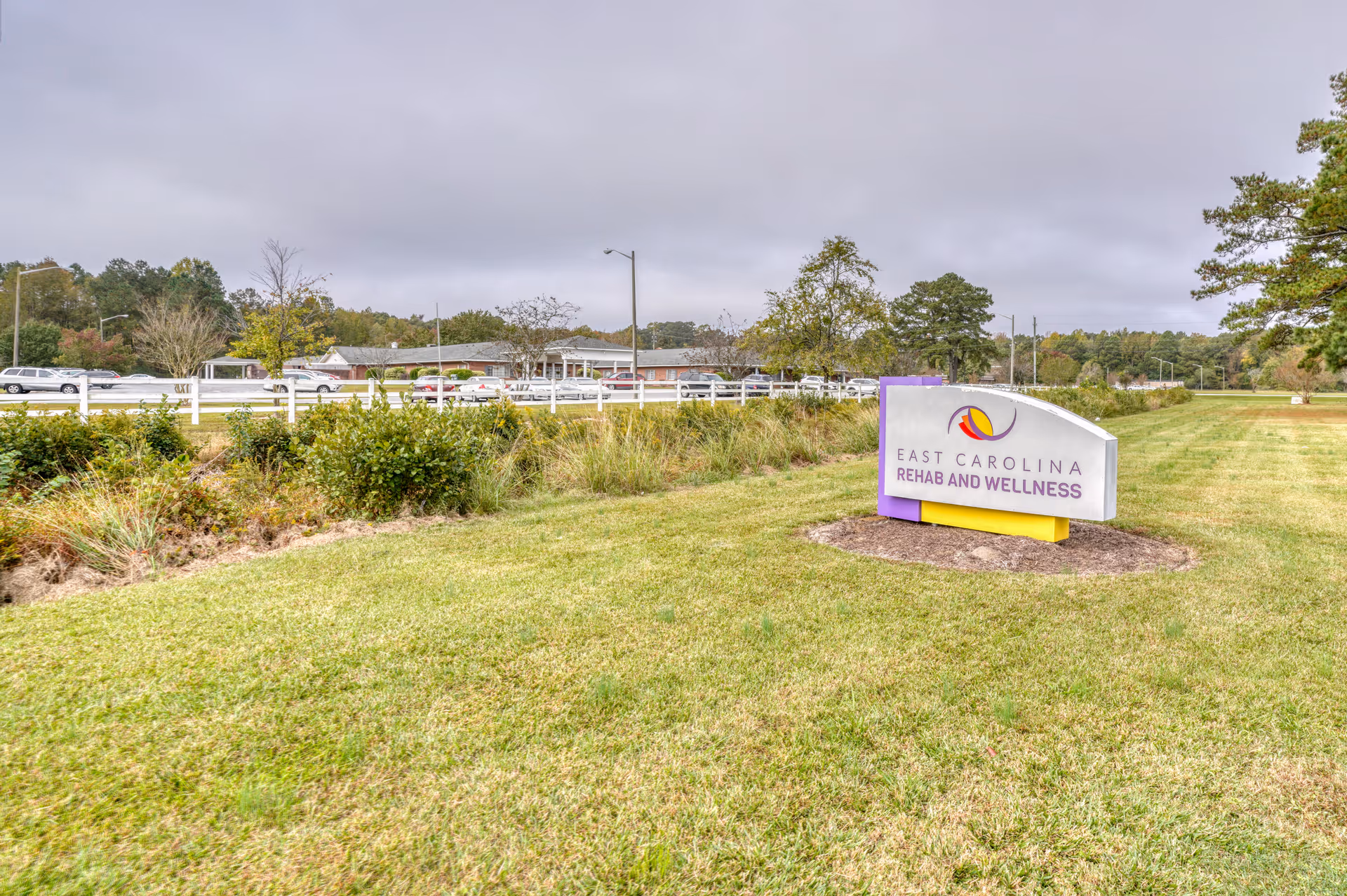 Outdoor view of East Carolina Rehab and Wellness facility sign on a grassy area with trees and a building in the background under a cloudy sky.
