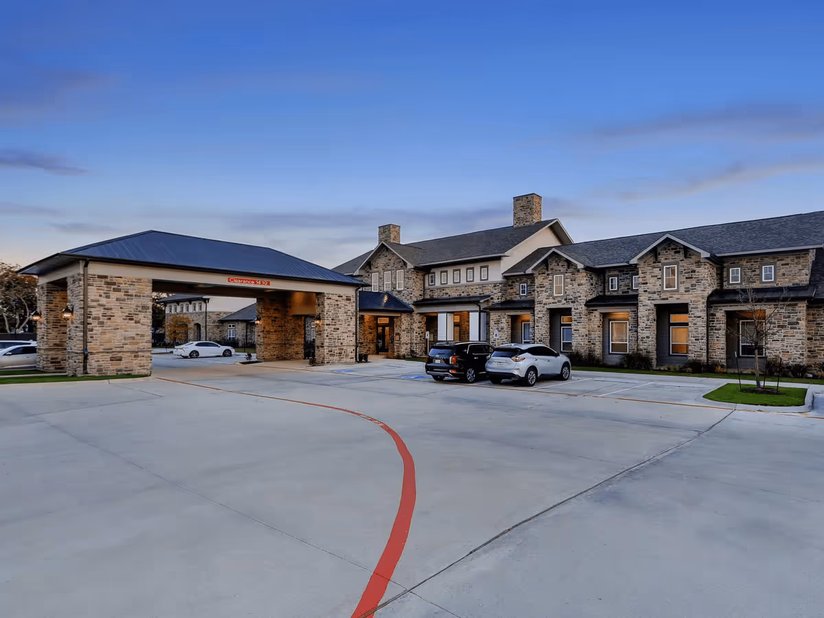 Stone-faced senior living building with a covered porte-cochere and parked cars in the front under a clear evening sky.
