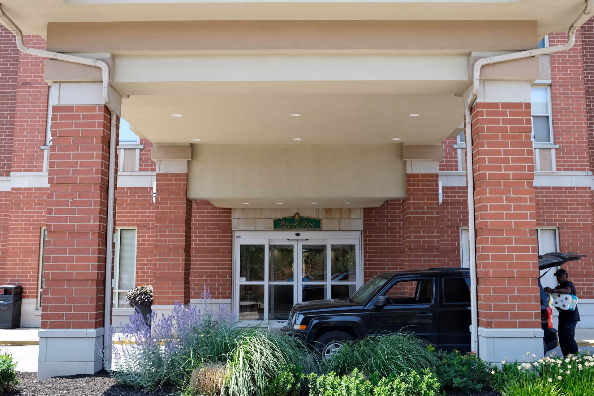 Entrance of a senior living facility with a covered drop-off area supported by brick columns. A black SUV is parked under the canopy, and a person is unloading items including a pack of paper towels from the vehicle. There are plants and flowers in front of the entrance, and a green sign above the doors reads 'Welcome Home A Senior Lifestyle Community'.